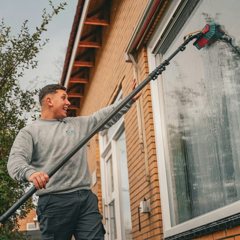 Mann reinigt Fenster mit einer langen Teleskopbürste vor einem Gebäude.
Glas- und Jalousienreinigung