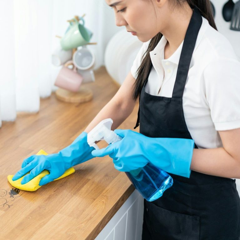 Frau mit blauen Handschuhen reinigt eine Arbeitsfläche mit einem Schwamm und Spray.
Unterhaltsreinigung